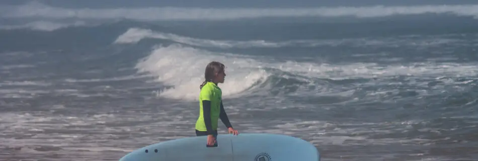 Young surfer in yellow rashguard entering ocean with blue soft top board